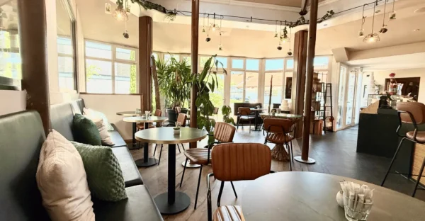 Gemütliche Sitzecke im Café der Tortenfee in Alling mit bequemen Kissen, stilvollen Holzstühlen, runden Cafétischen und grünen Zimmerpflanzen für ein einladendes Ambiente.