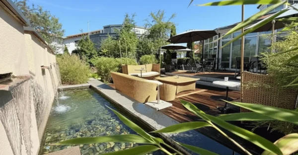 Sonnenterrasse des Cafés der Tortenfee in Alling mit modernen Lounge-Möbeln aus Holz, Sonnenschirmen und einem plätschernden Brunnen-Wasserlauf, umgeben von Bambuspflanzen.