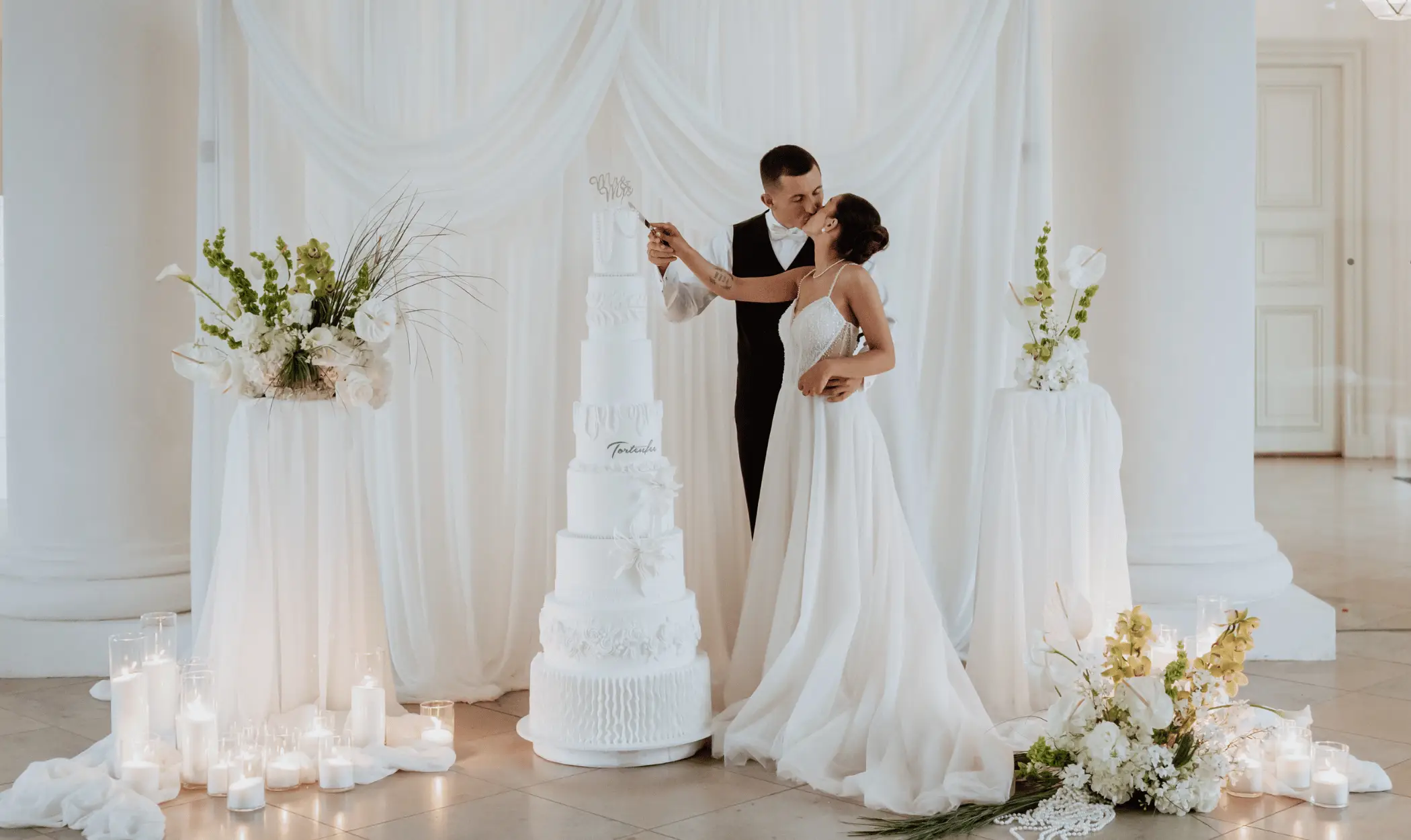 Ein frisch vermähltes Brautpaar schneidet küssend eine elegante, siebenstöckige weiße Hochzeitstorte von Tortenfee an, umgeben von floraler Dekoration und Kerzen in einer Münchner Hochzeitslocation.