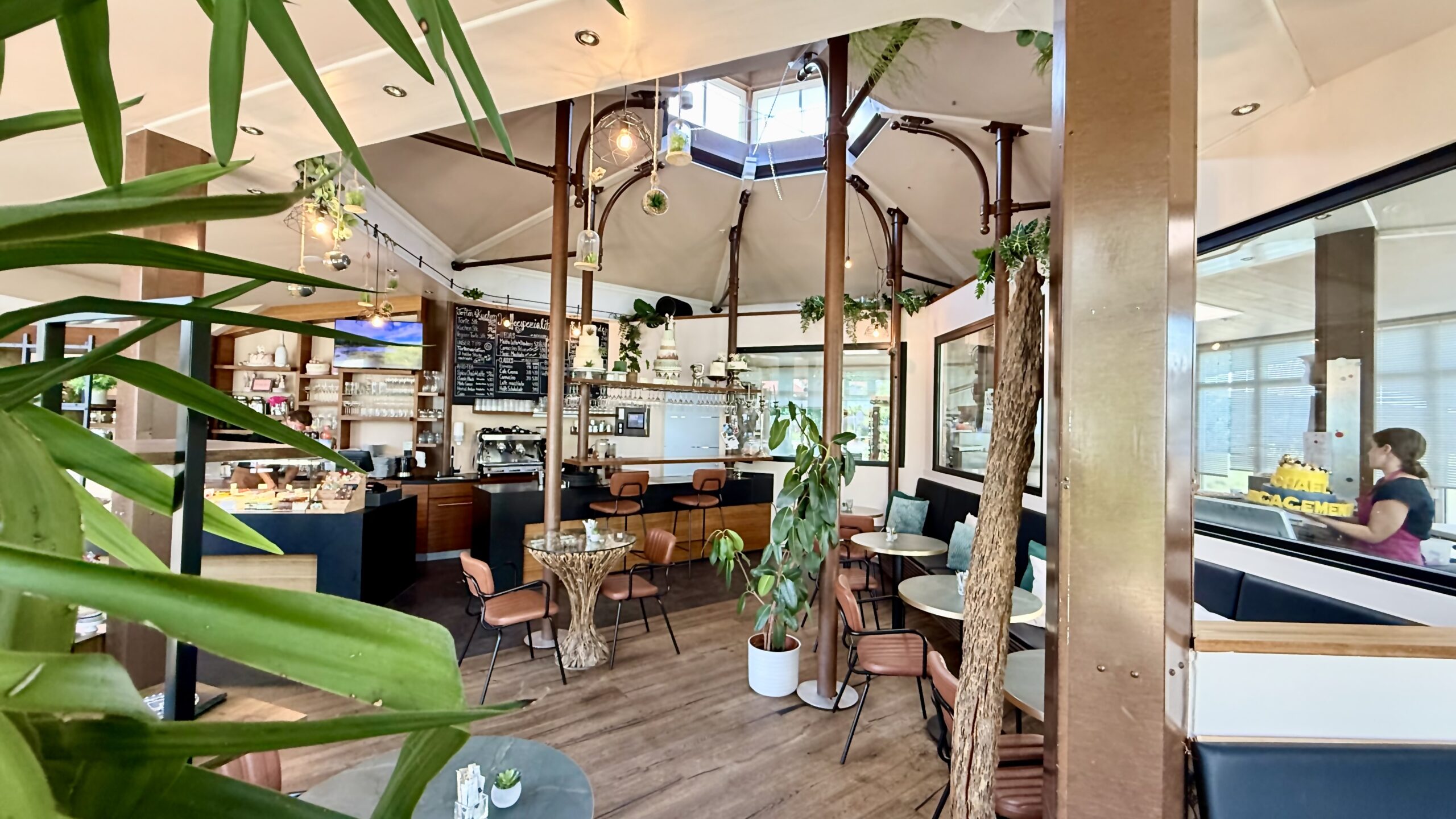 Gemütliche Sitzecke im Café der Tortenfee in Alling mit bequemen Kissen, stilvollen Holzstühlen, runden Cafétischen und grünen Zimmerpflanzen für ein einladendes Ambiente.