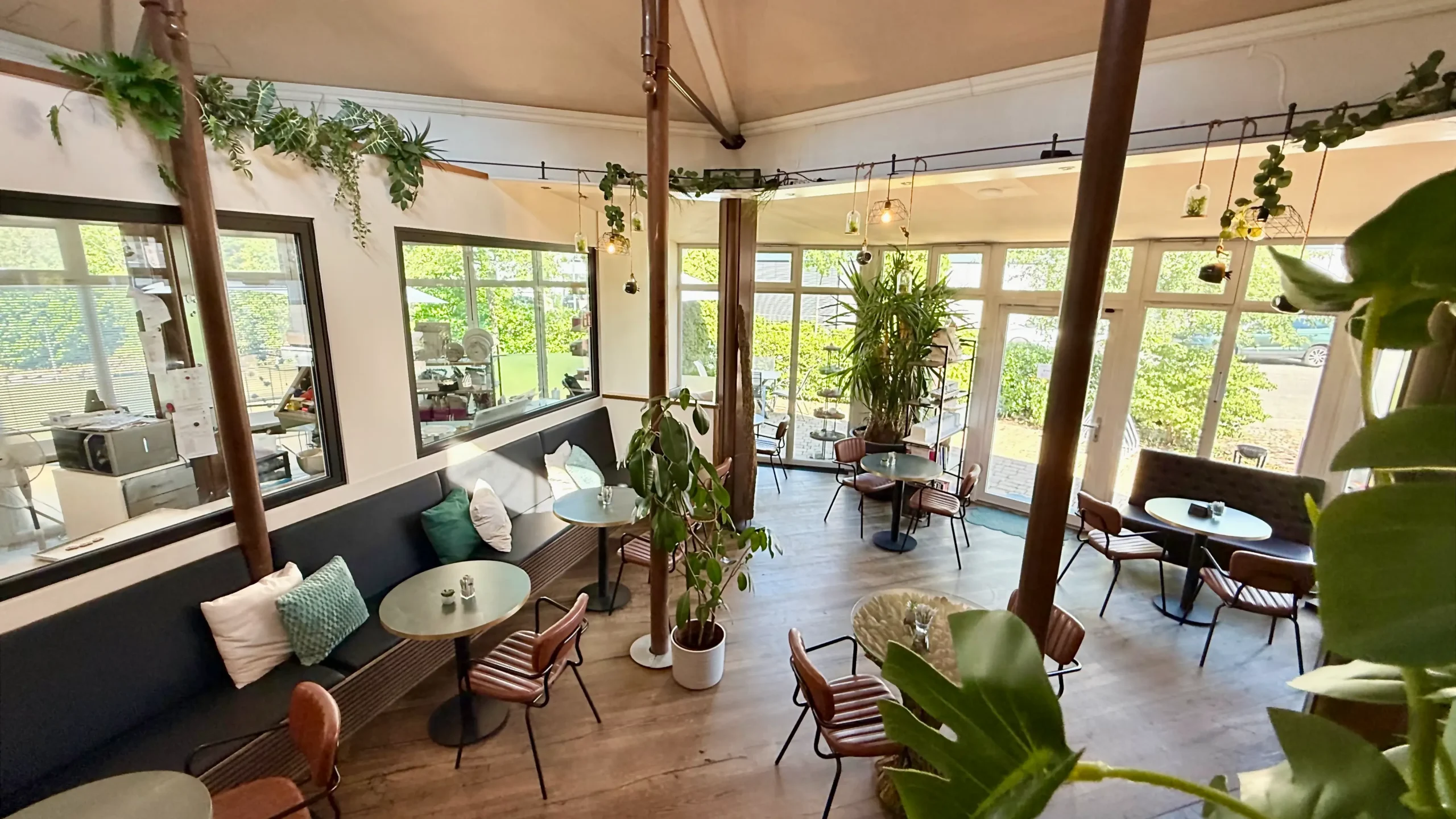 Moderner, lichtdurchfluteter Gastraum des Cafés der Tortenfee in Alling mit Blick auf gemütliche Sitzplätze, grüne Pflanzen und die gläserne Backstube im Hintergrund.
